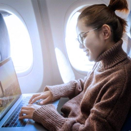 一位戴着眼镜、穿着棕色毛衣的女士正在飞机上通过无线上网用笔记本电脑工作