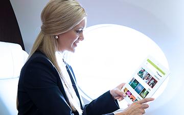 一个留着金色长发的女人坐在飞机上用平板电脑浏览Viasat网站