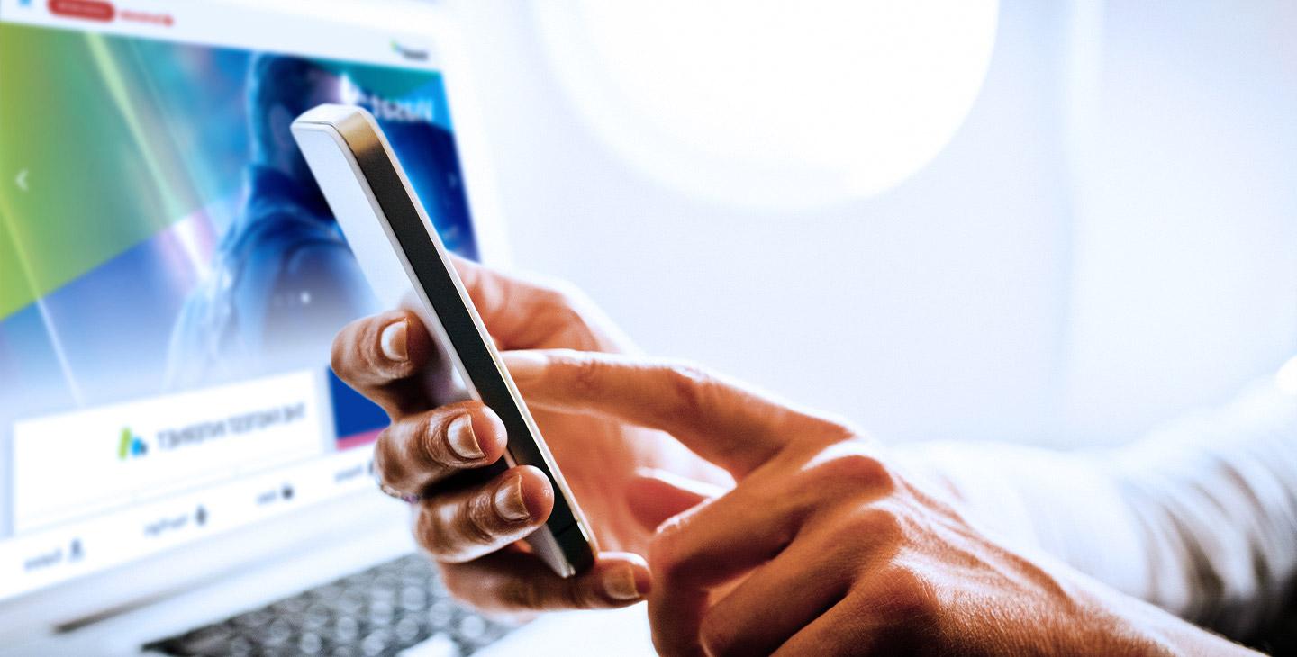 Close-up of a hands holding a smartphone wtih a laptop in the background showing inflight advertising of a movie