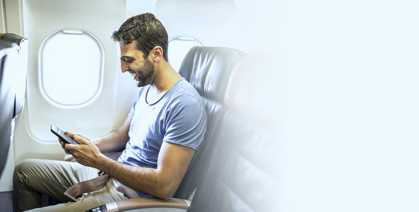 一名留着棕色头发和胡须的男子在飞机上坐在窗边看他的智能手机