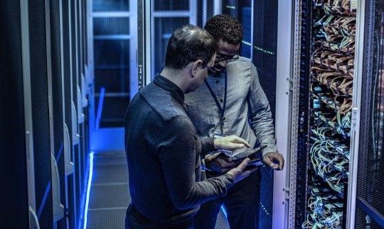 两个男人在服务器室讨论政府的网络安全，看着一台平板电脑