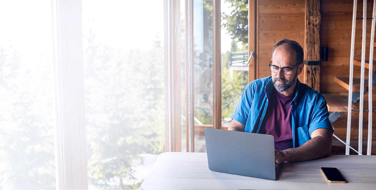 一个拥有家庭无线网络计划的男人坐在他山上的房子里的桌子前，手里拿着笔记本电脑 