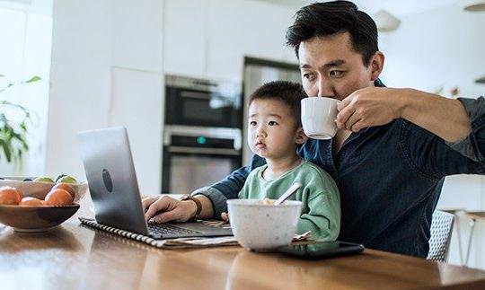 一个拥有家庭电话和互联网的男人坐在厨房的桌子旁，他的儿子坐在他的腿上, 看着笔记本电脑