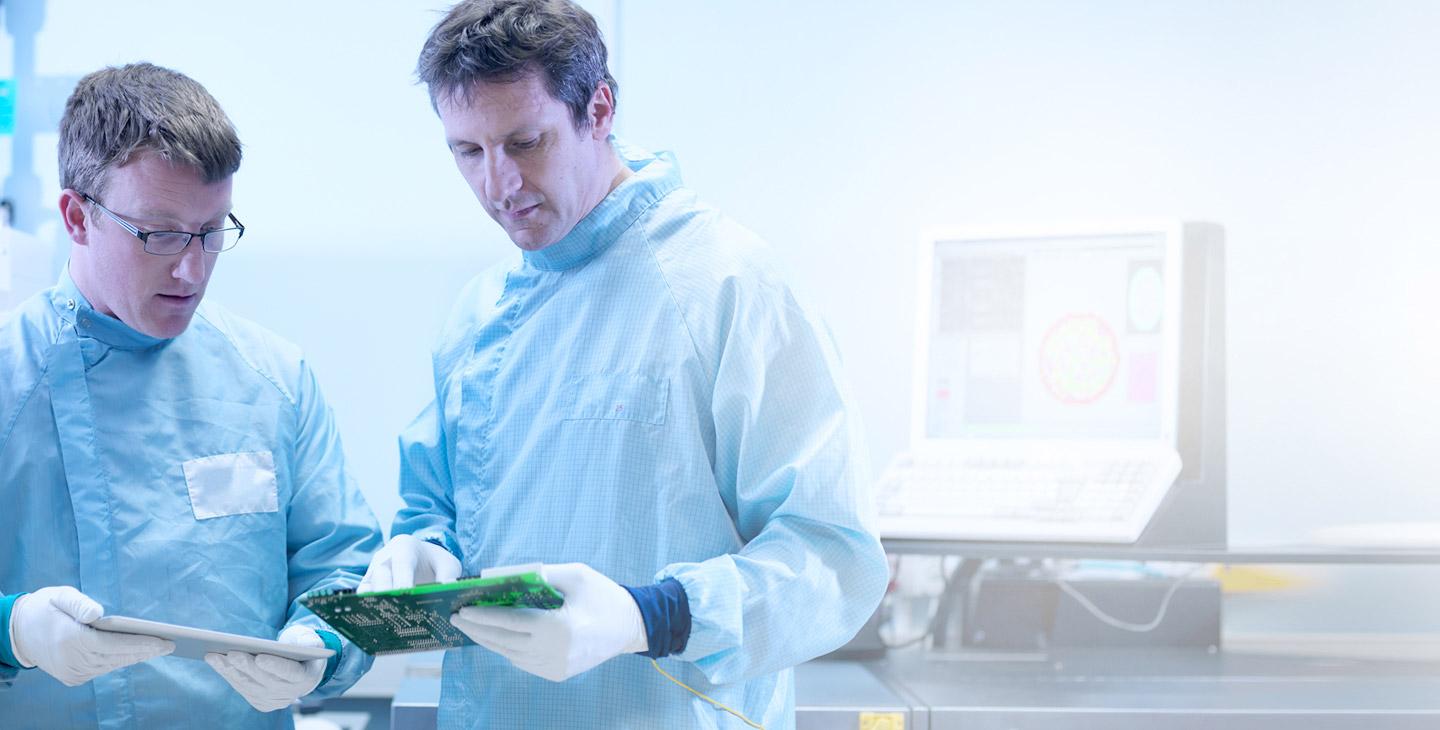 两个穿着实验室制服，戴着乳胶手套的人看着一个半导体，其中一个拿着一个平板电脑